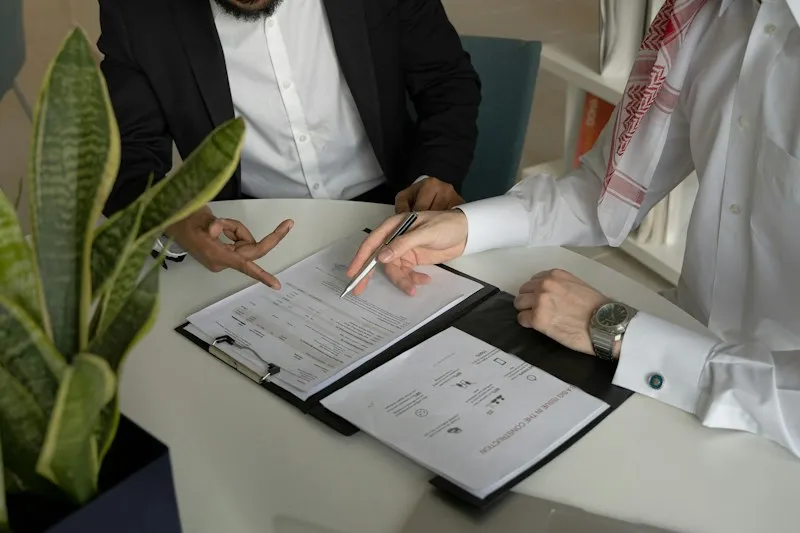 Two men at a table pointing at printed documents with a pen, one in a white thobe and red-checked shemagh