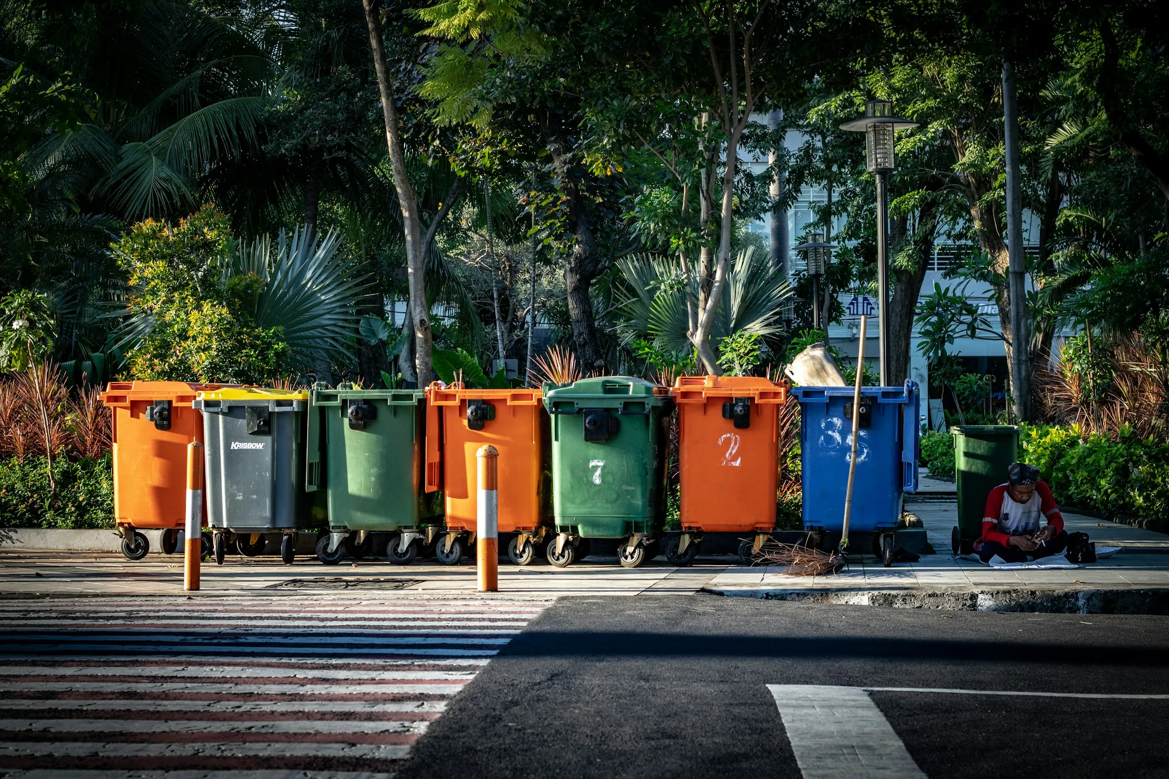 A row of orange, green, grey and blue wheeled waste containers lines a city pavement in warm sunlight.