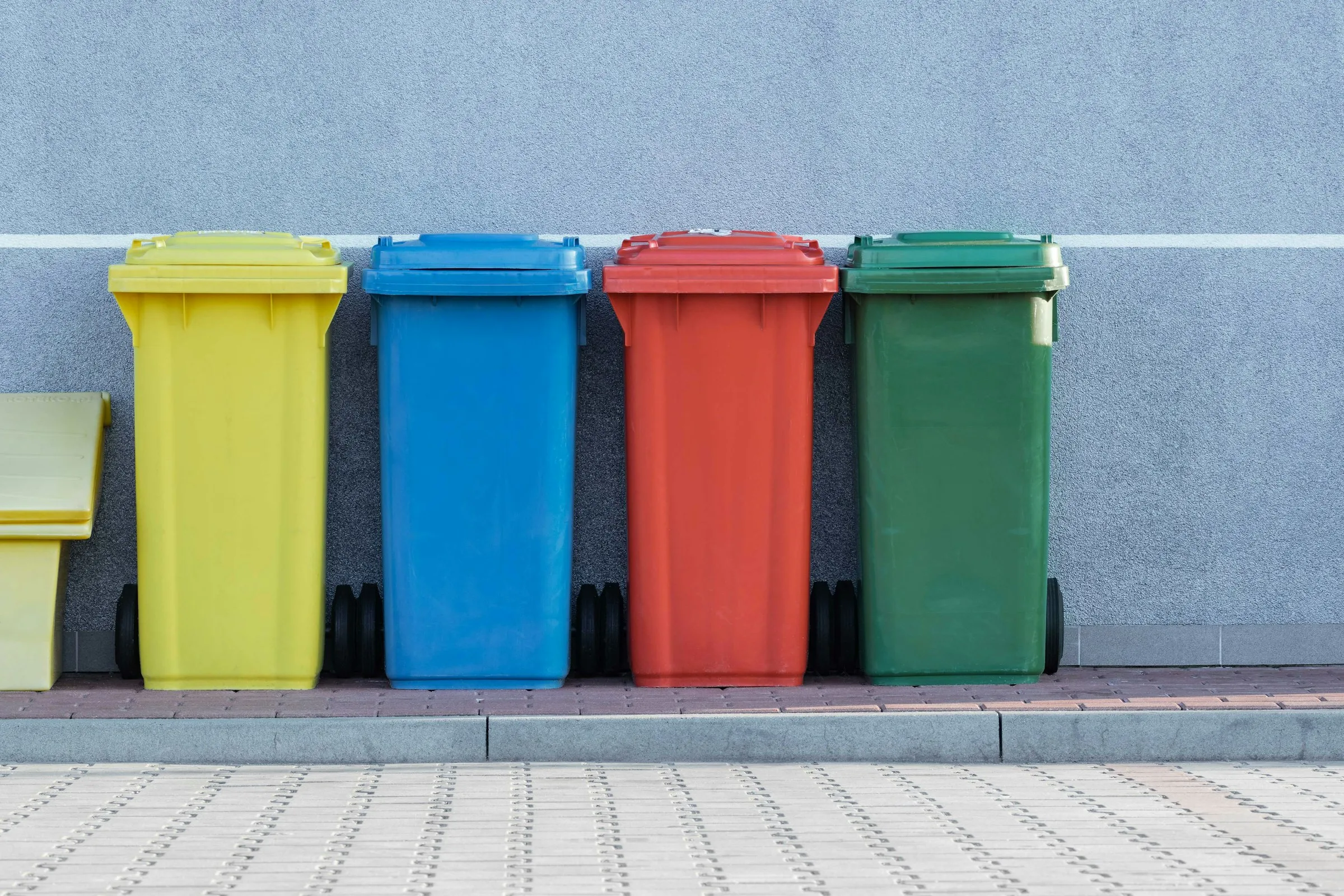Four wheelie bins in yellow, blue, red and green stand in a line against a grey wall.