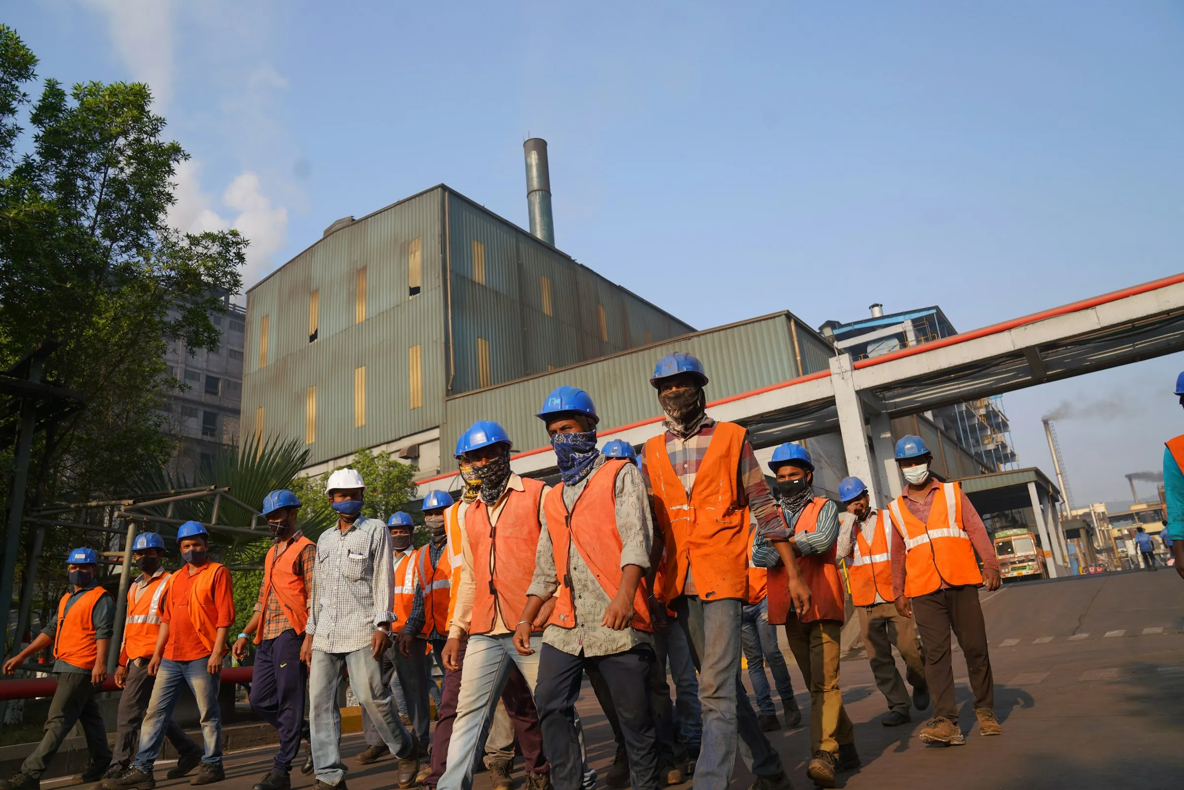 A crew in orange hi-vis vests and blue hard hats walks together through an industrial plant on a sunny day.