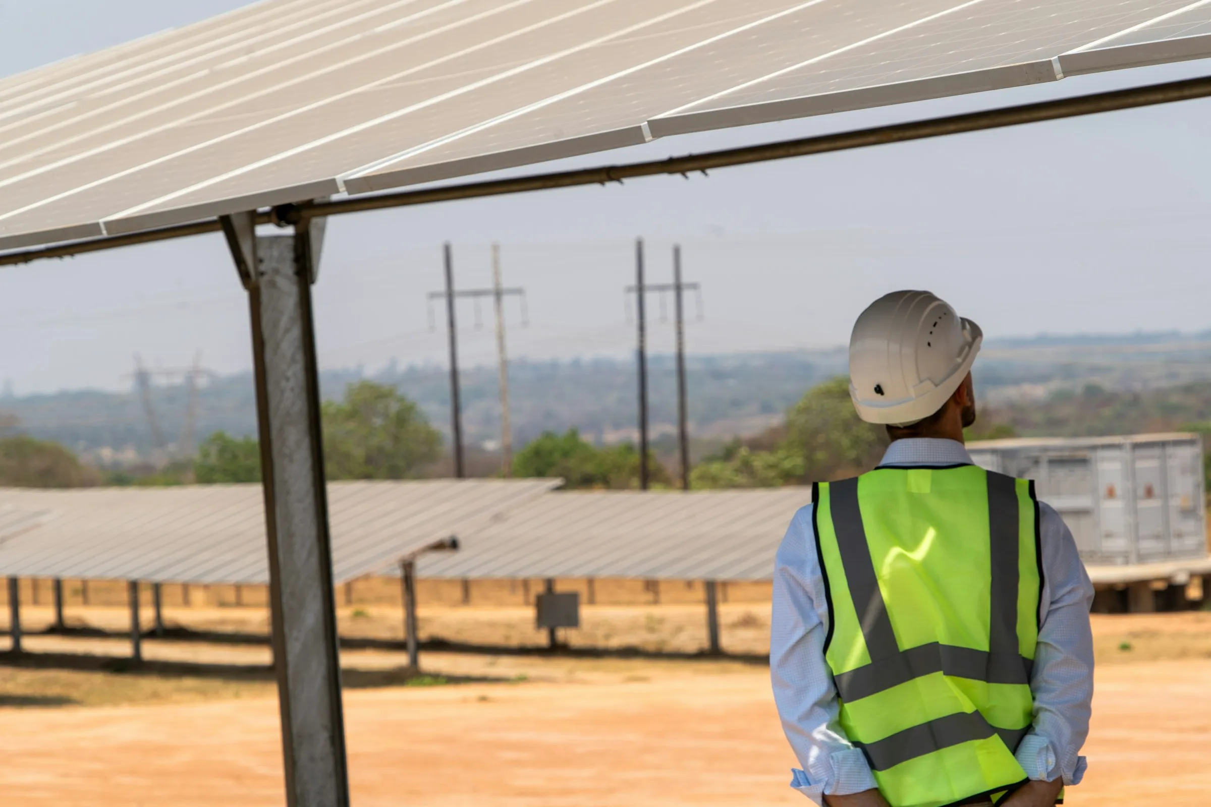 An inspector in a white hard hat and hi-vis vest looks over a solar installation on dry, open ground.