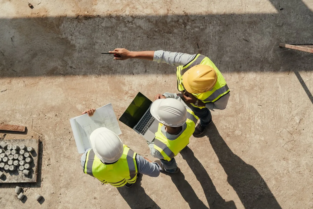 Three construction workers wearing helmets and high-visibility vests reviewing blueprints and a laptop on a construction site.