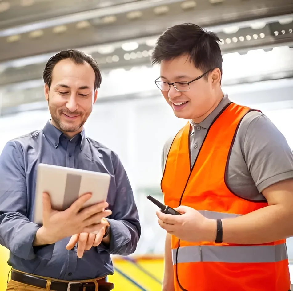 A manager holding a tablet and a smiling worker in an orange high-visibility vest holding a handheld device, looking at the screen together in a bright industrial building.