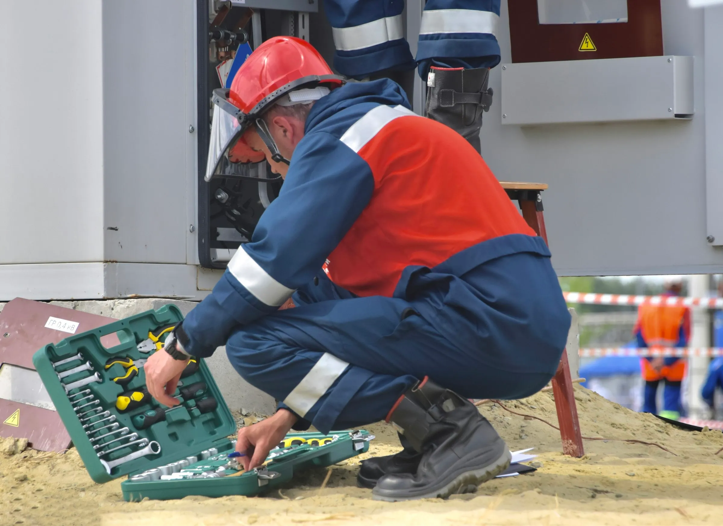 A technician in a hard hat and face shield crouches over an open toolkit beside an electrical cabinet.
