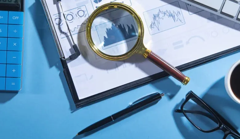 Magnifying glass over financial charts on a clipboard, with a pen, calculator, eyeglasses, and coffee cup on a blue desk.