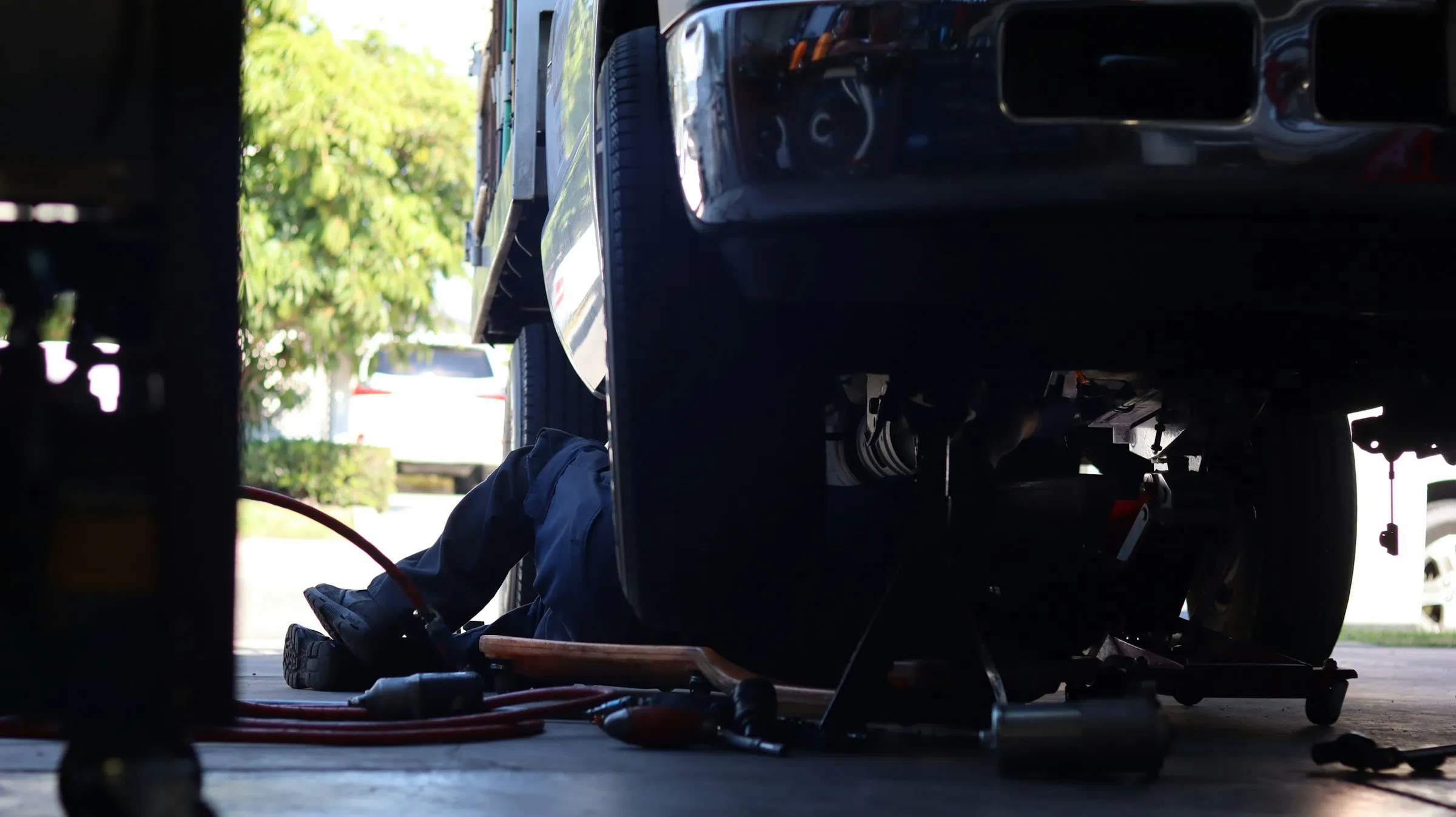 A mechanic lies under a raised vehicle in a workshop with tools and an air hose on the floor.