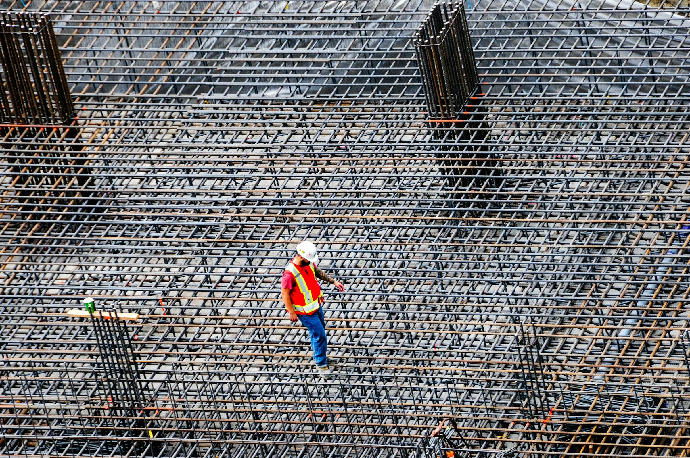 A construction worker in a hard hat and hi-vis vest walks across a large grid of steel rebar, checking the reinforcement before the concrete is poured.