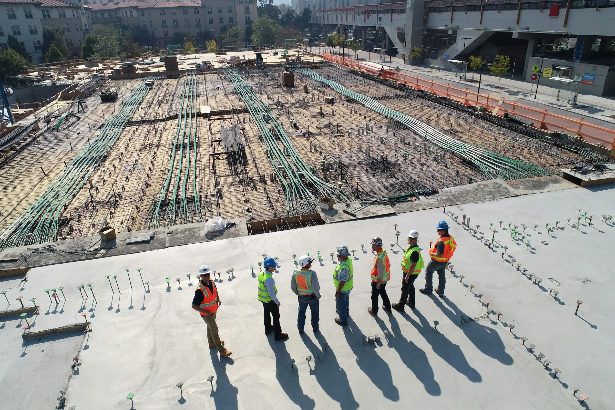 Seven construction workers in hard hats and hi-vis vests stand in a line on a concrete deck, seen from above.