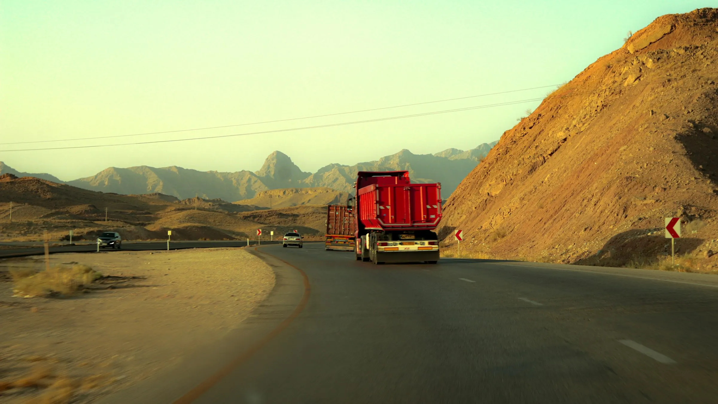 A red truck drives along a desert highway towards rocky mountains in warm evening light.