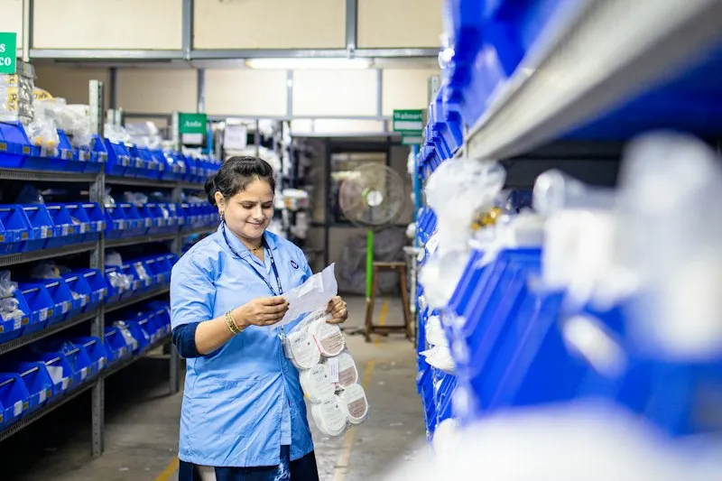 A storeroom worker in a blue uniform checking a paper list against stock on shelves of blue parts bins