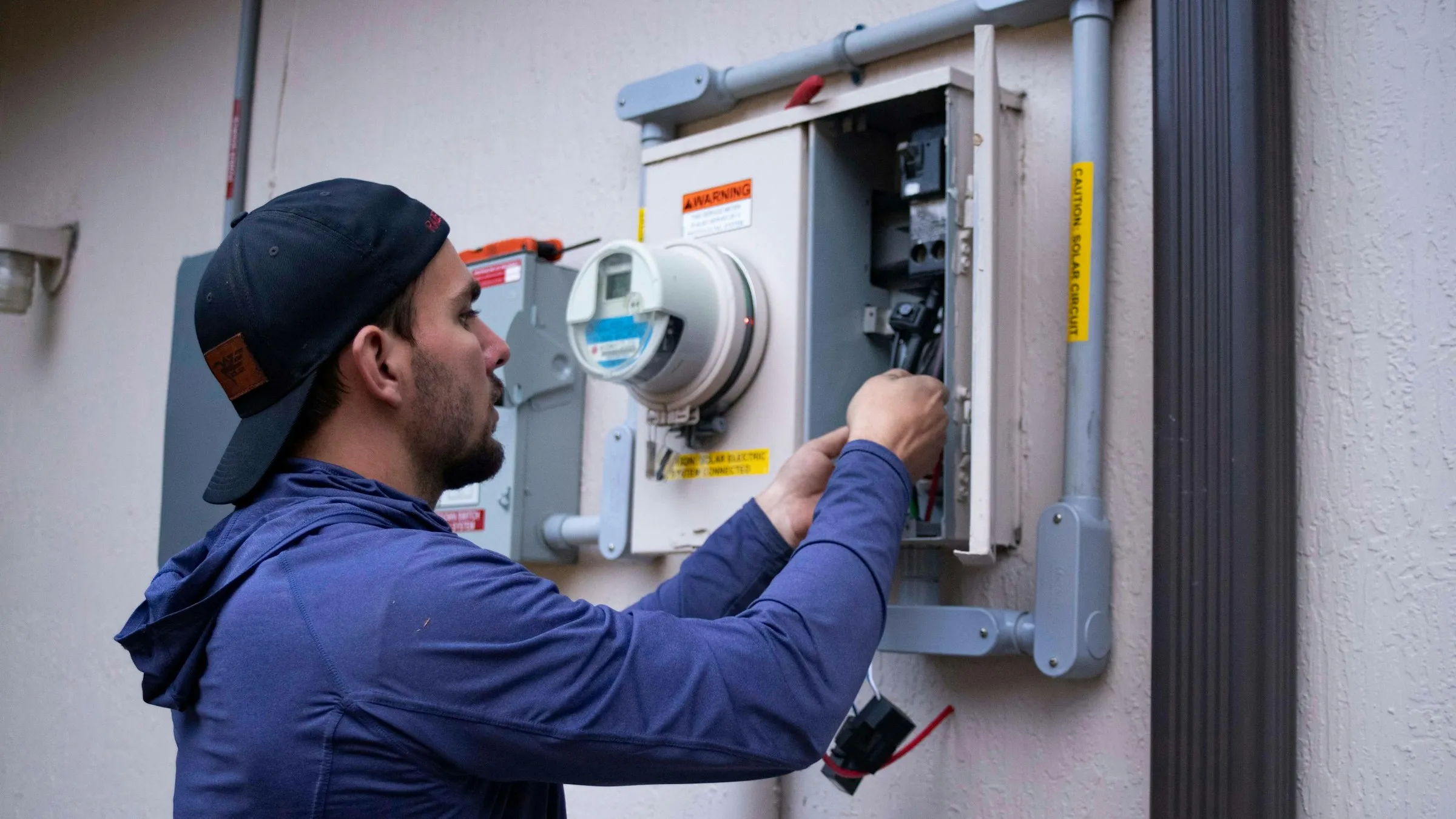 A technician wires up an electricity meter cabinet mounted on a pale stucco wall.