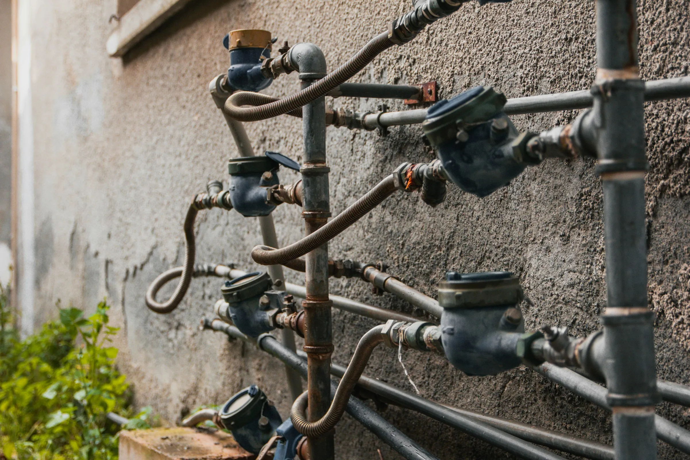 A row of water meters connected by pipes and flexible hoses runs along a weathered concrete wall.