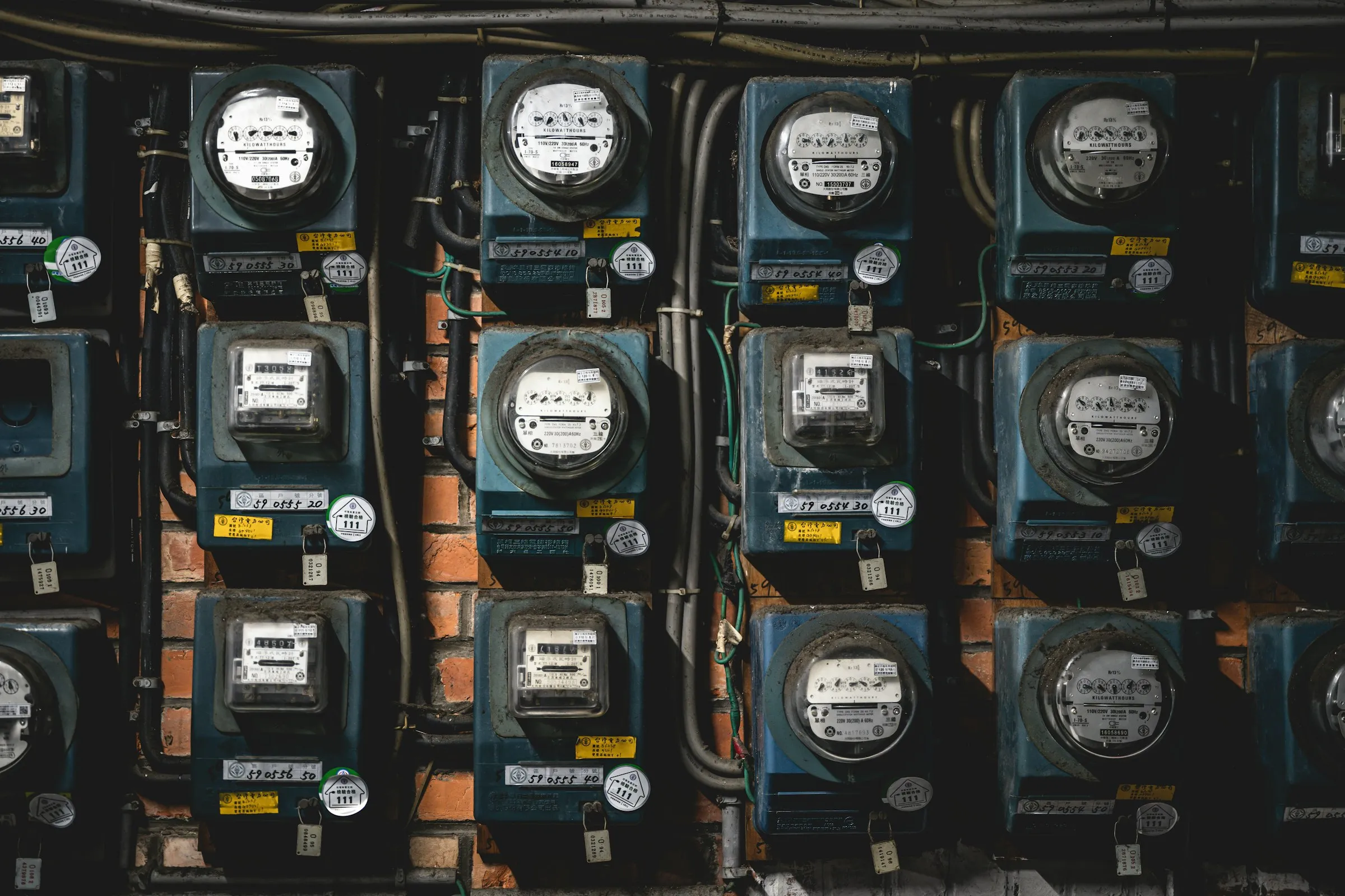 Rows of tagged electricity meters fill a brick wall, each with its own dial and sealed cover.