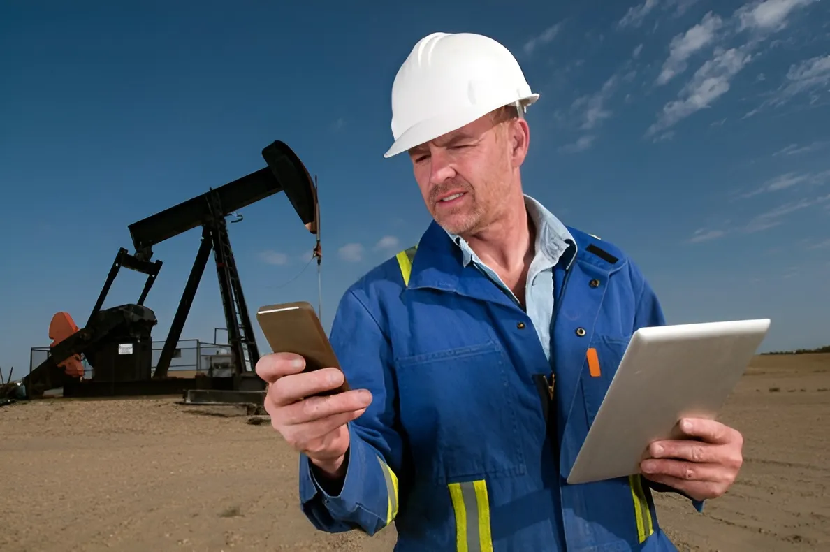 Oil field worker in a hard hat using a phone and tablet with a pump jack behind him