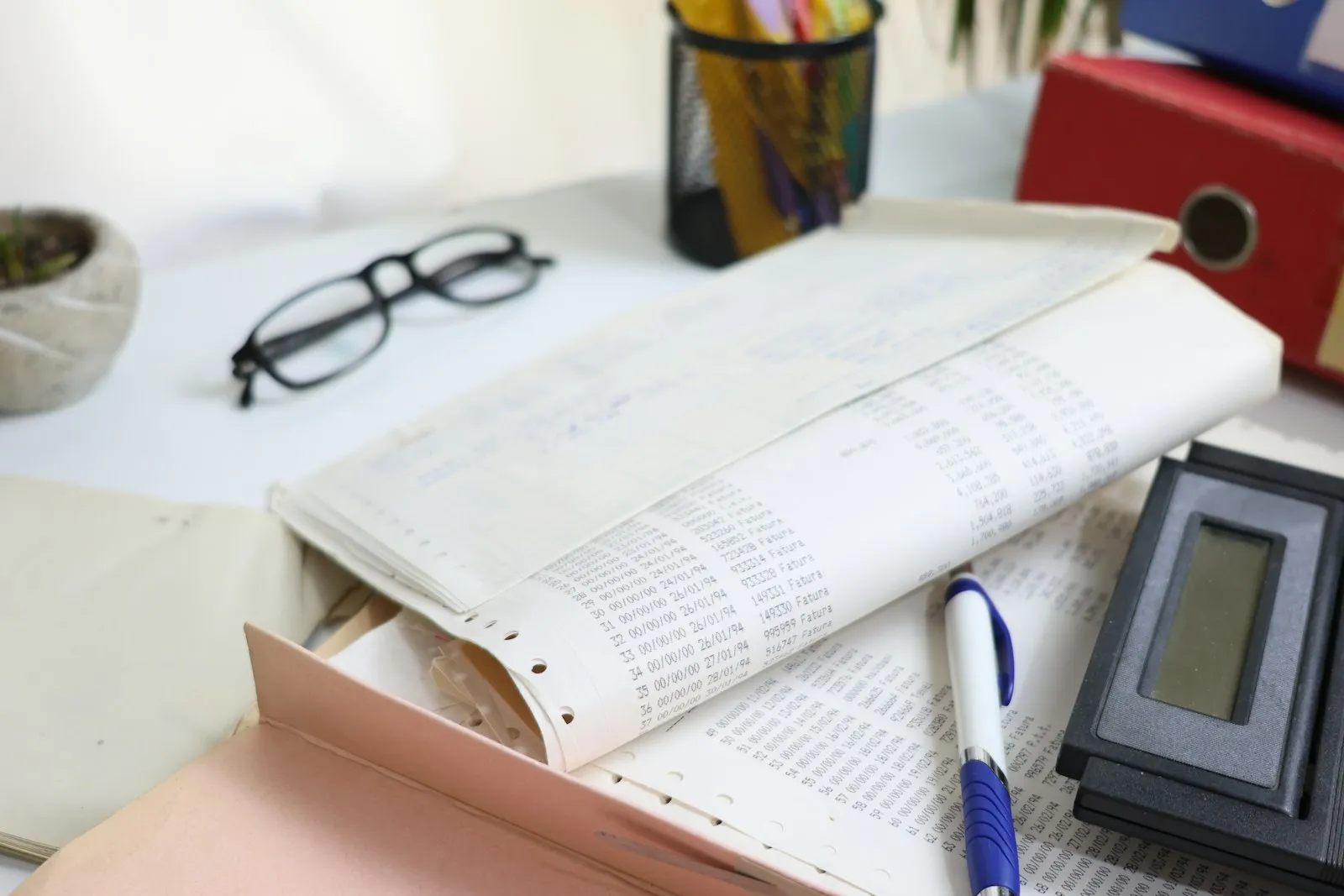 A desk covered with printed ledger sheets, with a calculator, glasses and a pen