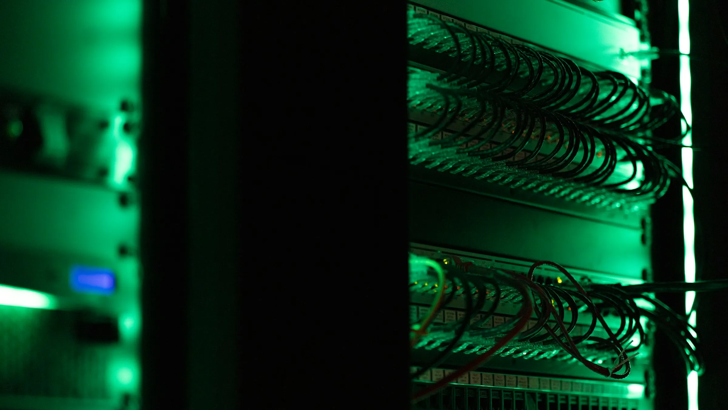 Server racks full of neatly bundled network cables glow softly green in a dark server room.