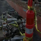 A pile of broken bricks beside a brick wall with torn yellow caution tape sagging from an orange traffic cone.