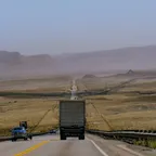 A box truck driving away along a long, straight highway through dry hills with few other vehicles in sight.