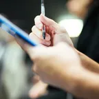 A close view of a hand holding a pen over a blue clipboard, filling in a paper form.