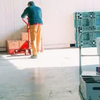 A worker pulls a red pallet jack stacked with cardboard boxes across a bright warehouse floor, with empty pallets and a trolley nearby.