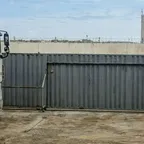 A white truck cab stopped beside a closed grey corrugated metal sliding gate with barbed wire on the wall behind it.