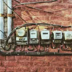 A row of five old electricity meters mounted side by side on a red brick wall, with cables running between them.