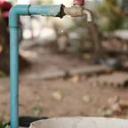A worn blue water pipe and tap drips a single drop of water above a paint-stained bucket on a dirt floor.