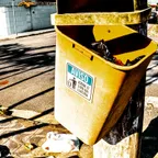 A yellow street bin knocked loose and tilted on its post, with rubbish bags and litter spread on the pavement below.
