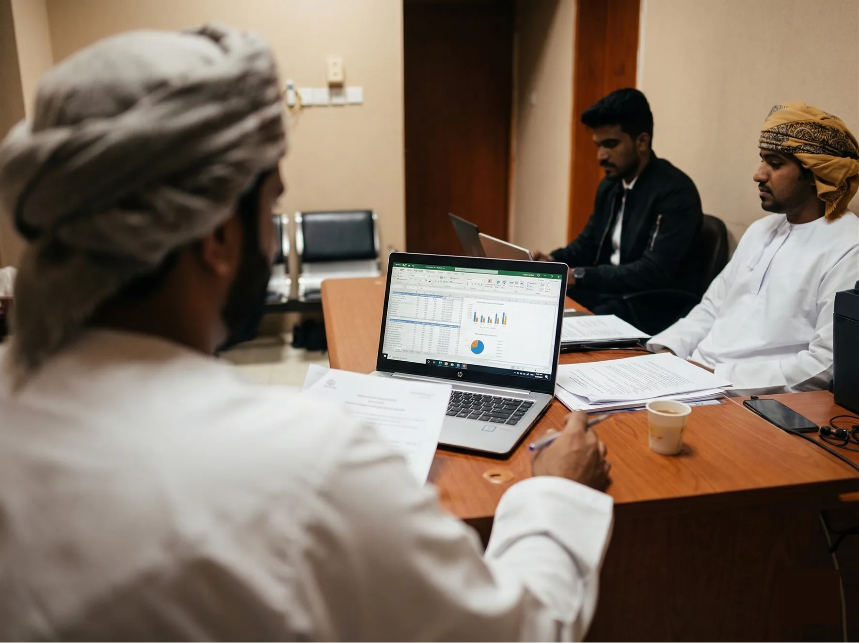 Three men in an office meeting room working on laptops and reviewing paper documents at a wooden table