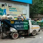 A small municipal rubbish truck piled high with bags of waste, parked on a street in front of a painted wall.
