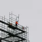 A worker in an orange jacket and white hard hat stands on a high steel scaffold platform against a grey sky.