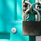 A dark padlock and rusty chain fastened across a bright turquoise metal door.