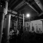 A black-and-white view of a dim basement plant room with insulated pipes, valves and a single bare bulb overhead.