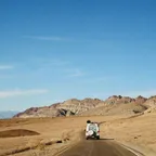 A white 4x4 van driving alone along a narrow road through empty desert with rocky hills ahead.