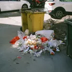A pile of rubbish bags and food packaging spilled on a sunlit street beside two yellow wheeled bins, with parked cars behind.