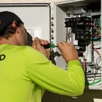 A technician in a lime-green work shirt and cap uses a screwdriver on the wiring inside an open electrical box on a wall.