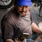 A mechanic in a blue cap and grease-stained hands looks at a smartphone while sitting beside motorbikes in a workshop.