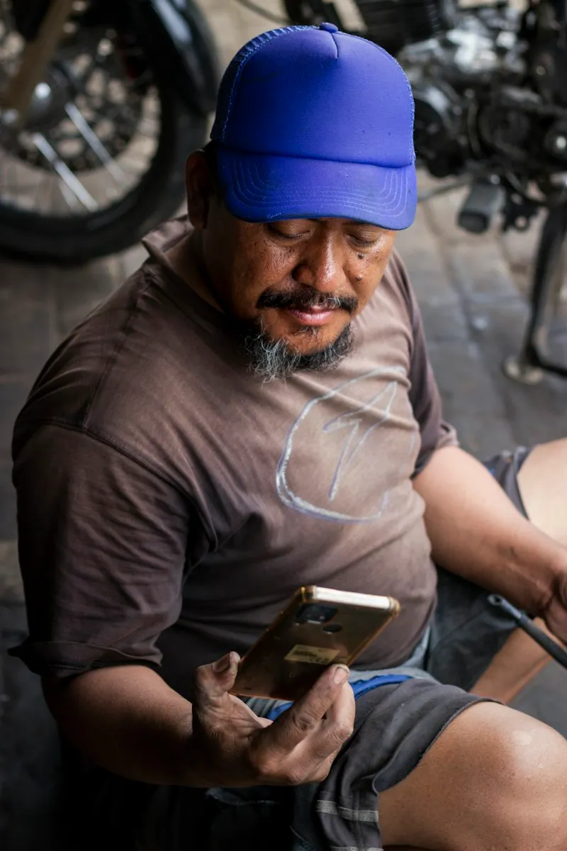 A mechanic in a blue cap and grease-stained hands looks at a smartphone while sitting beside motorbikes in a workshop.