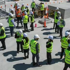 A group of people in hard hats and hi-vis vests gathered in a circle on a concrete rooftop at a construction site.