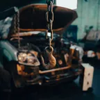 A vehicle with its hood raised and engine exposed sits in a dim workshop behind a hanging hoist chain and hook.