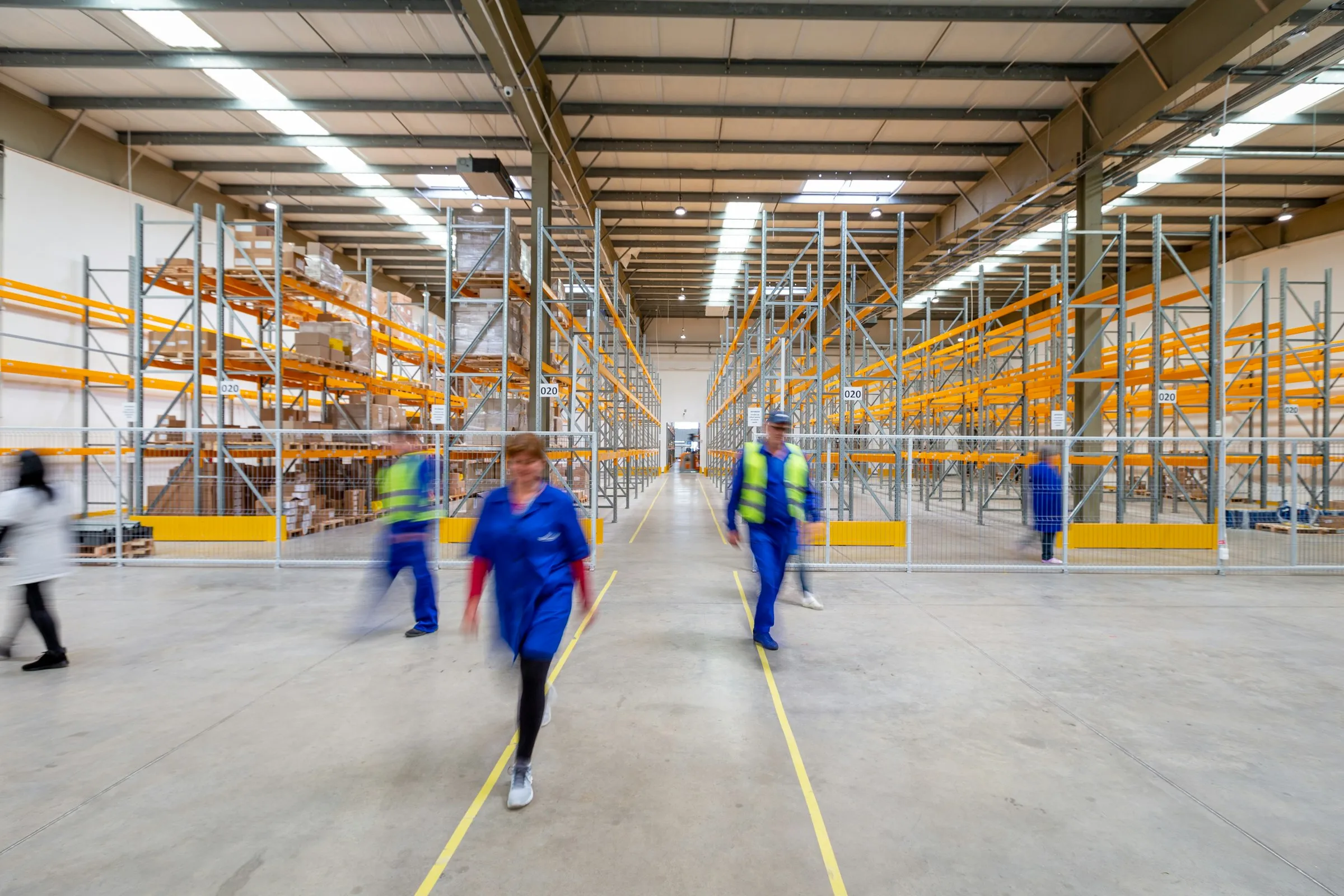 Warehouse staff in work clothes and hi-vis vests walk along an aisle between tall orange pallet racks.