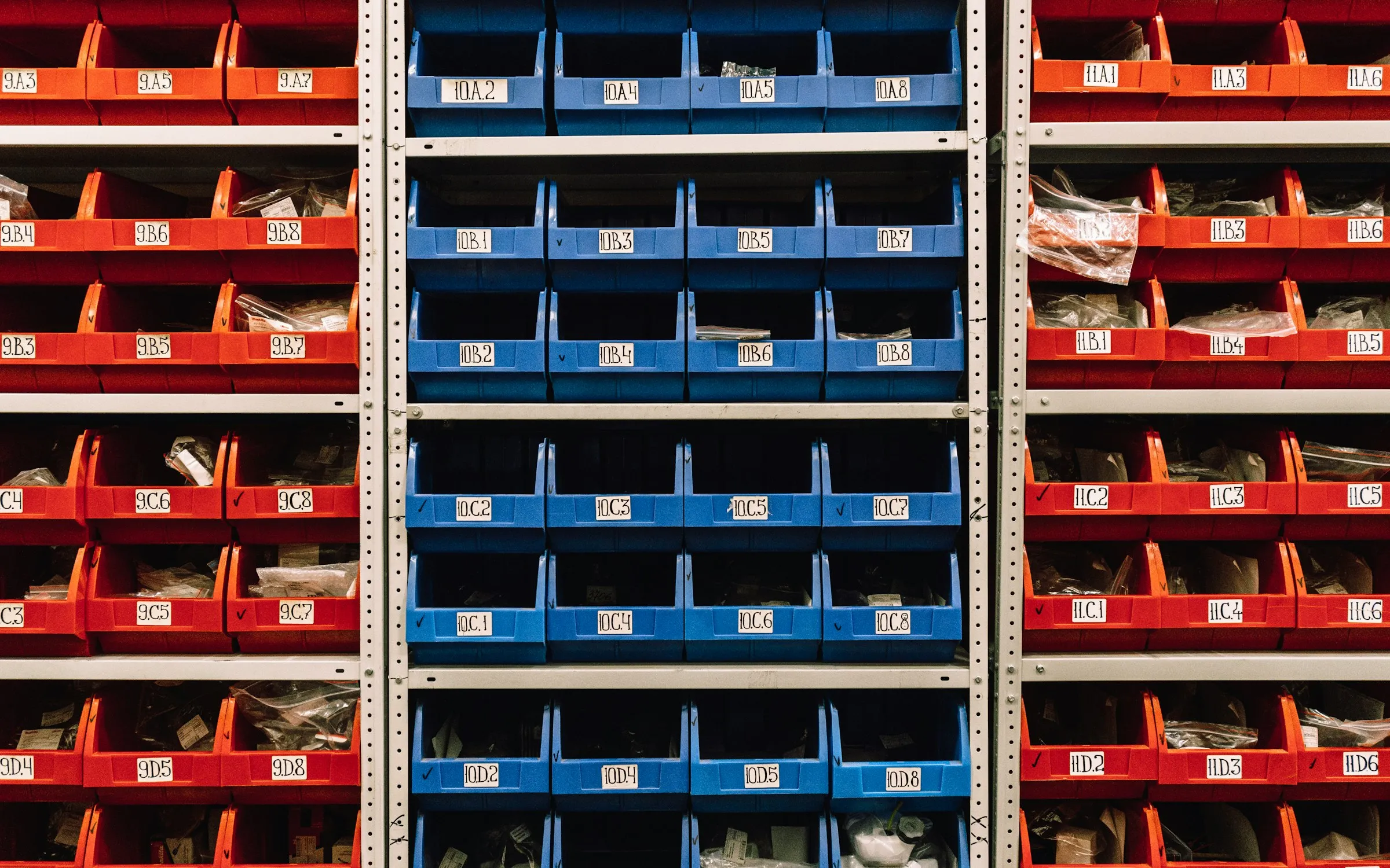 A wall of red and blue parts bins on metal shelving, each bin labelled with its own location code.