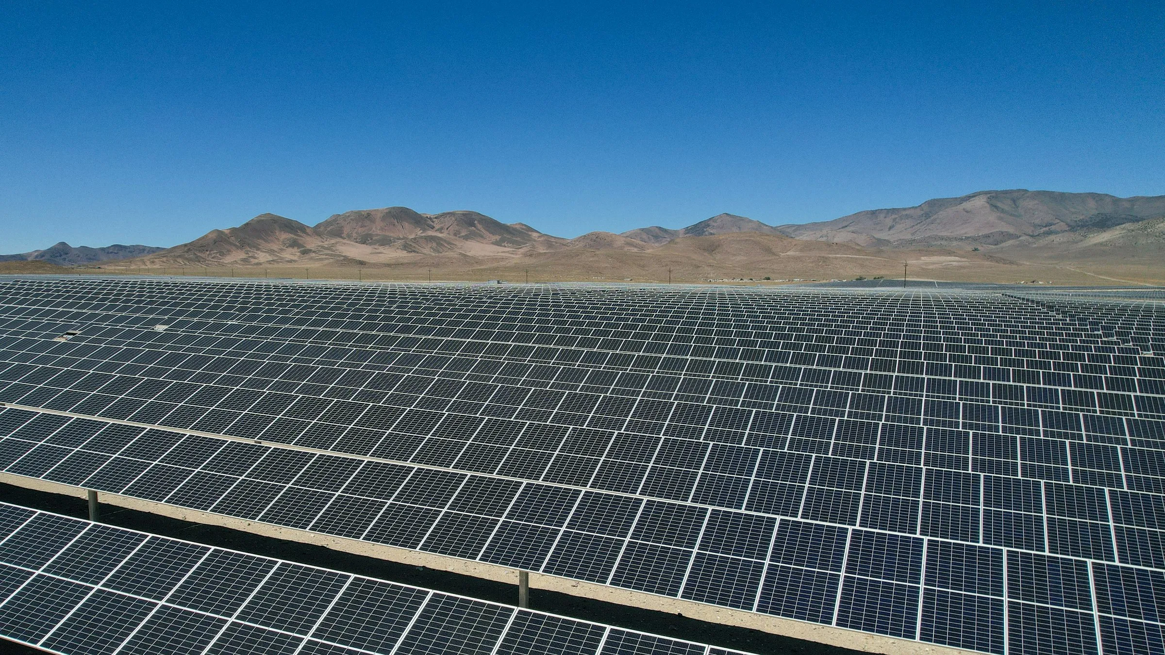 Rows of solar panels stretch across a dry valley toward bare desert mountains under a clear blue sky.