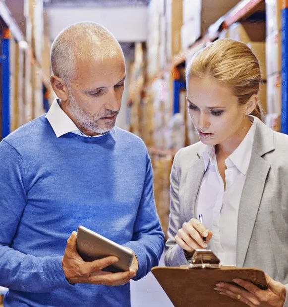 Two colleagues in a warehouse aisle checking stock on a tablet and a clipboard