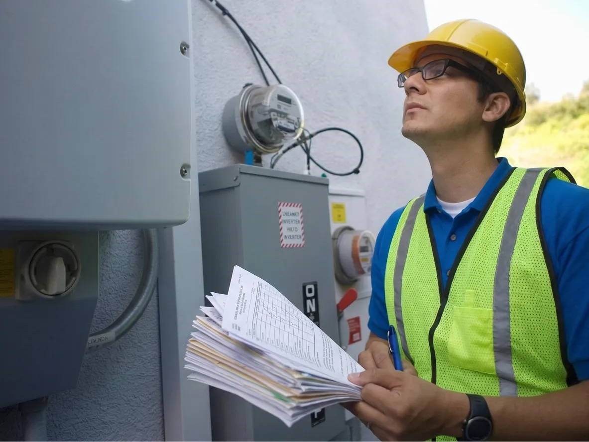 Worker in a hard hat and safety vest holding a stack of paper forms next to electrical equipment