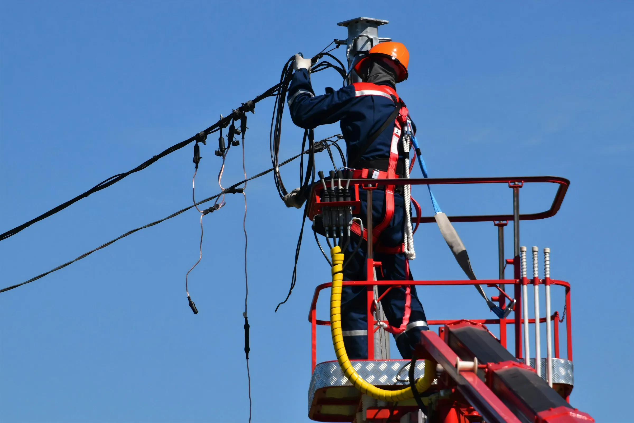 A line worker in a hard hat and safety harness repairs overhead power cables from a raised lift platform.